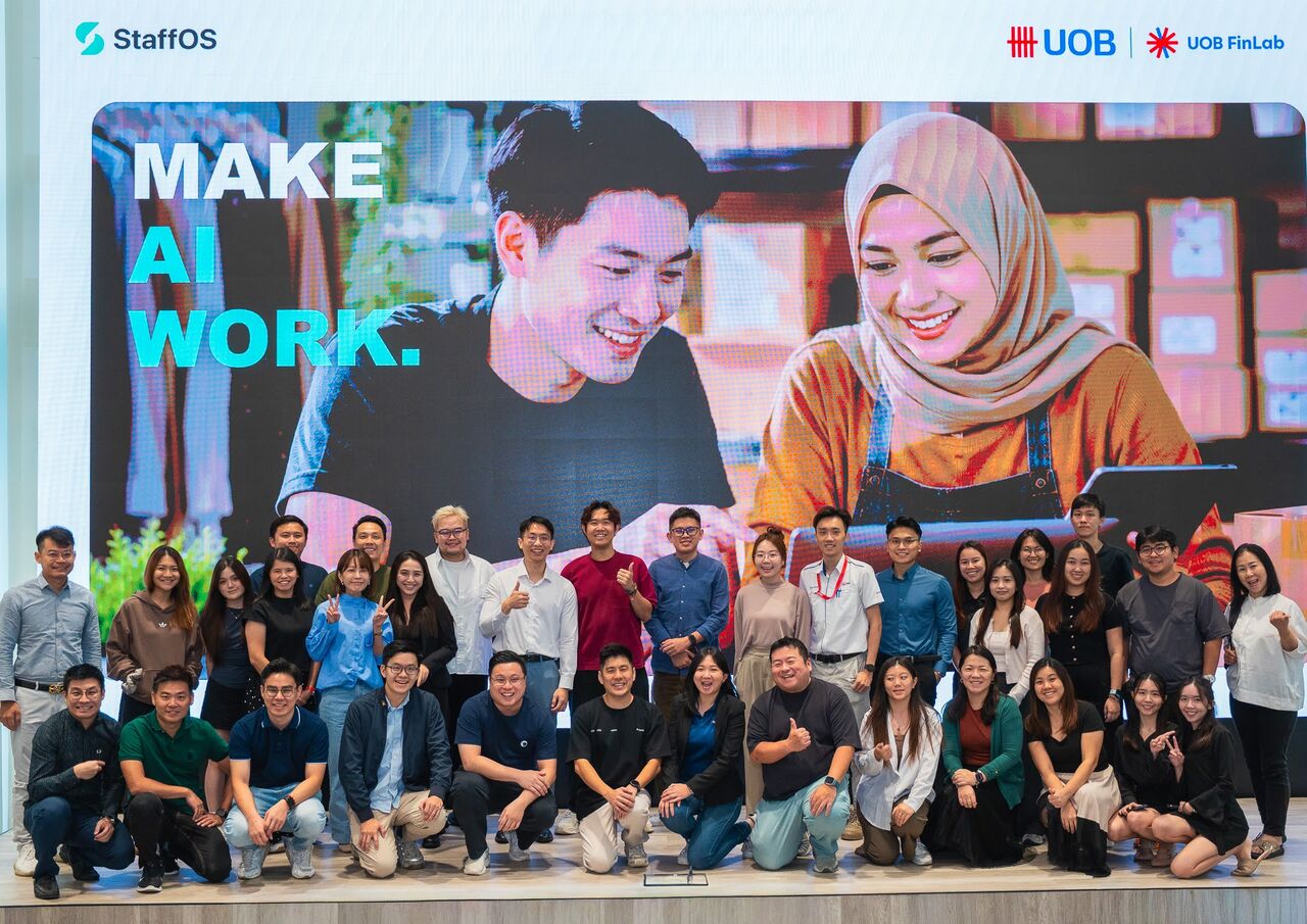 Attendees and hosts of the Make AI Work workshop pose for a group photo on stage, in front of a screen with the StaffOS and UOB FinLab logos.